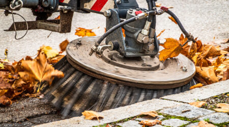 Side view of a street sweeper cleaning a sidewalk with dead leaves all around