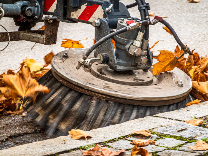 Side view of a street sweeper cleaning a sidewalk with dead leaves all around